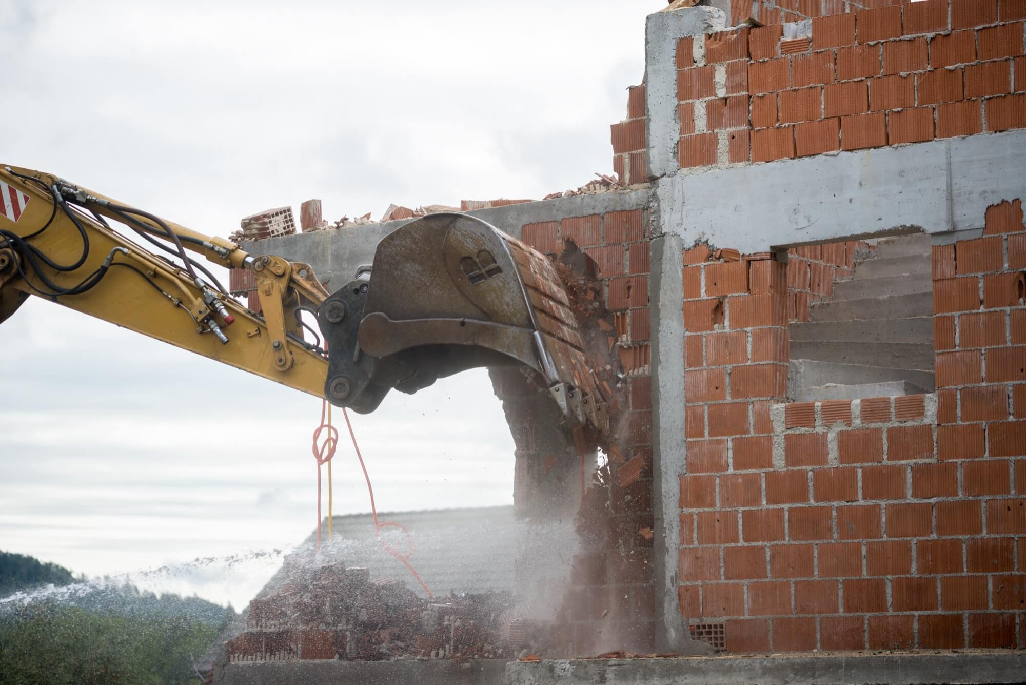 A yellow excavator performing demolition of a brick wall. Water is sprayed to control the dust from the demolition work.